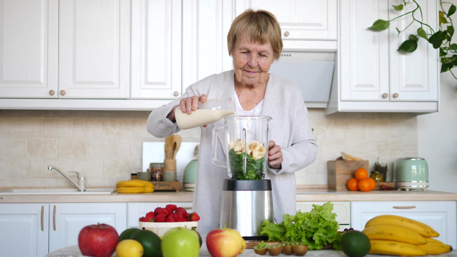 Ältere Dame steht in der Küche und bereitet sich einen Smoothie zu.