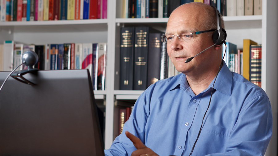 Mann sitzt mit Headset vor einem Bildschirm, im Hintergrund ein gefülltes Bücherregal