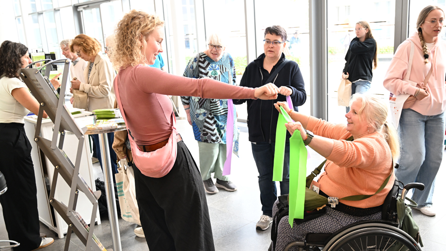 Frau im Rollstuhl zieht ein Theraband zwischen den Händen, eine junge Frau steht davor