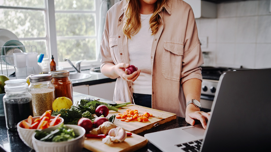 Junge Frau schneidet Gemüse, neben ihr geöffneter Laptop, ihr Gesicht ist nicht zu sehen