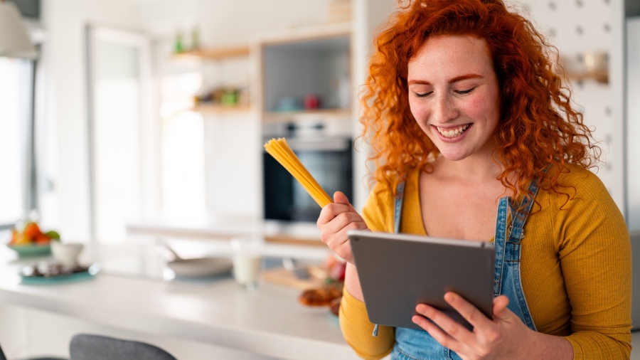 Junge Frau in einer Küche, hält Spaghetti und ein Tablet in den Händen
