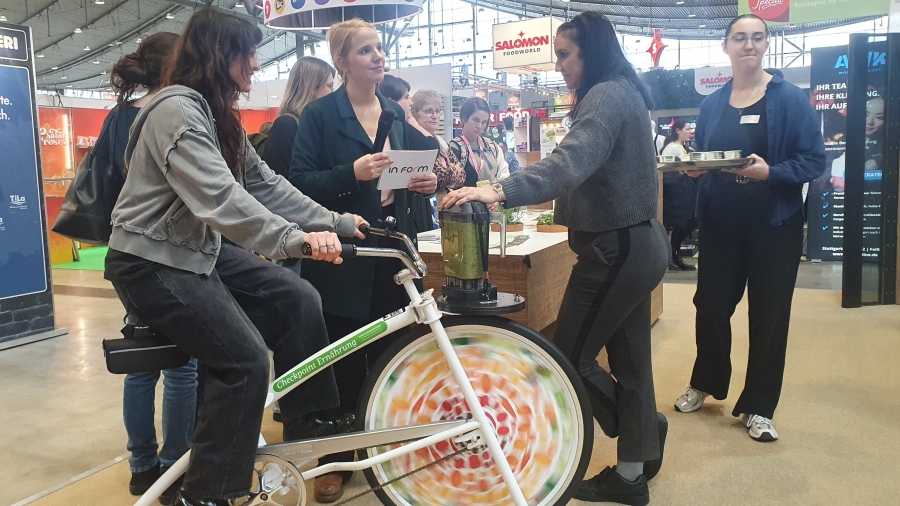Eine Frau auf dem Smoothie-Bike, zwei Frauen stehen daneben (Messestand)