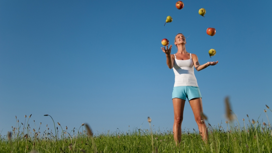 Frau steht auf einer Wiese und jongliert mit 6 Äpfeln