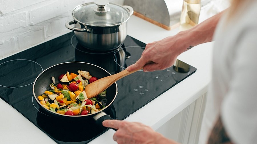 Auf einer Herdplatte wird Gemüse in einer Pfanne gebraten. Daneben steht ein geschlossener Kochtopf.