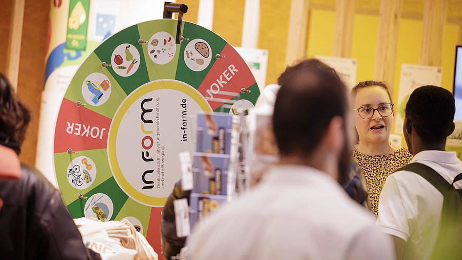 Glücksrad IN FORM, daneben Frau mit Brille und zwei junge Männer