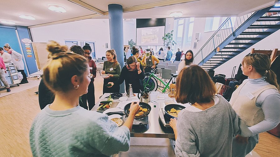 Mehrere Menschen stehen um einen Tisch herum, auf dem gekocht wird und unterhalten sich.