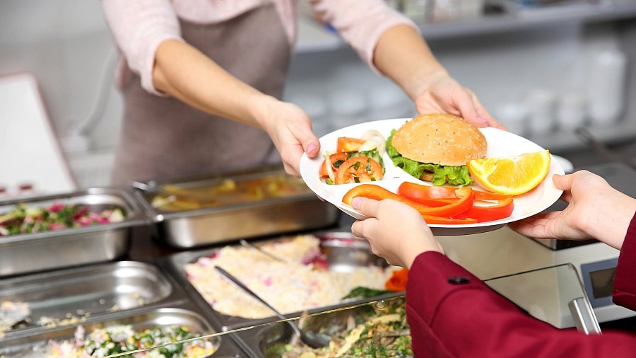 Küchenkraft reicht einem Kind ein Tablett mit Essen in einer Kantine (Ausschnitt - Hände und Essenstablett)