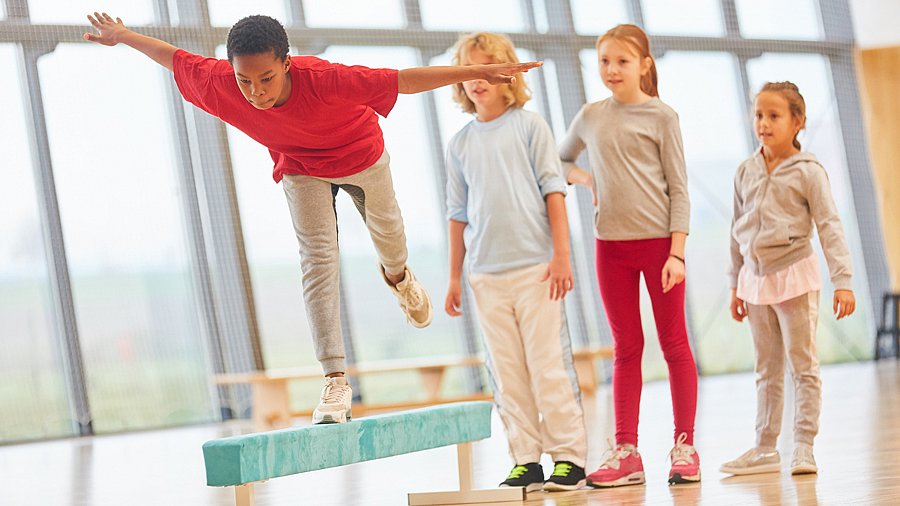 Vier Kinder im Turnunterricht, eines balanciert auf einem Schwebebalken