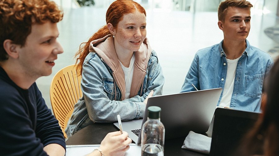 Drei Jugendliche, ein Mädchen und zwei Jungs, sitzen mit einem Laptop und weiteren Unterlagen an einem Tisch und sprechen mit anderen, in dem Ausschnitt nicht sichtbaren Personen.
