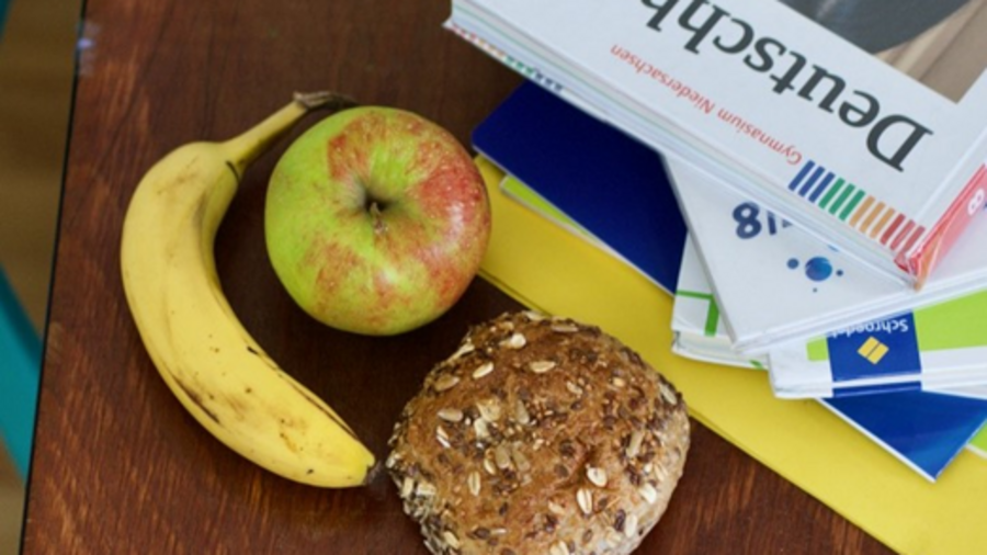 Auf einem Schultisch liegen ein Brötchen, eine Banane und ein Apfel neben Schulbüchern.