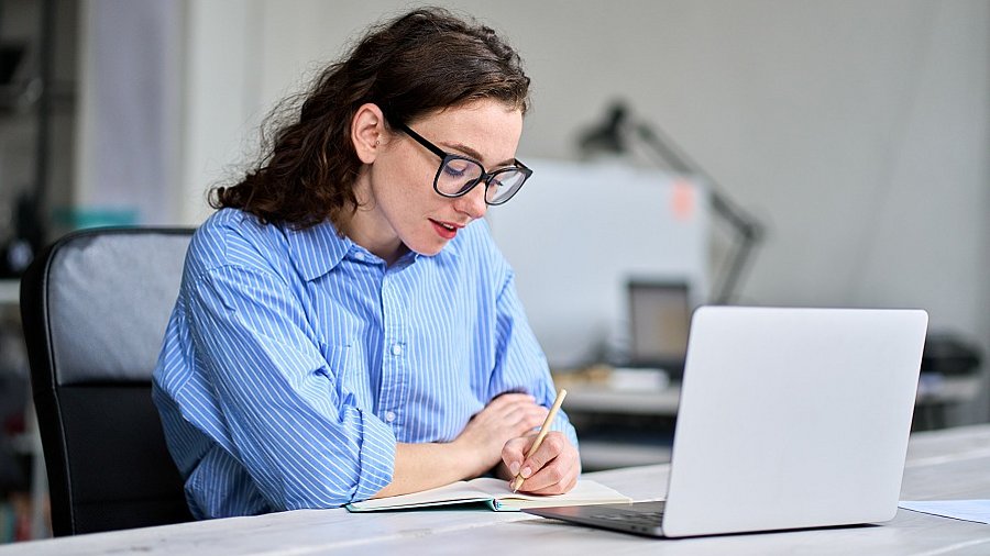 Frau sitzt vor Laptop und macht sich auf einem Block Notizen.