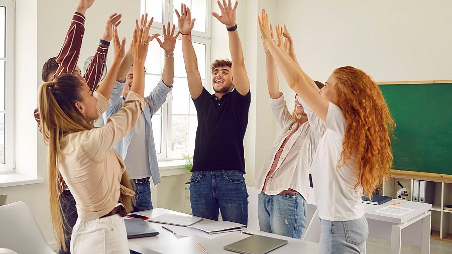 Gruppe Schüler steht um einen Tisch und hebt die Arme.