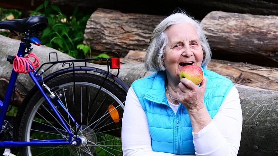 Fröhliche ältere Frau hält einen Apfel in der Hand, im Hintergrund steht ein Fahrrad