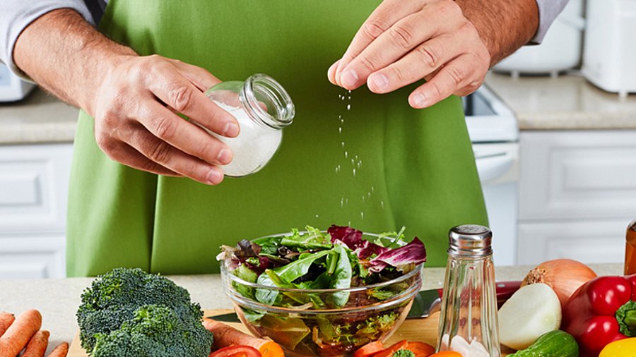 Hände streuen Salz aus einem Salzstreuer über einen Salat, darum eine Auswahl an verschiedenen Gemüsesorten