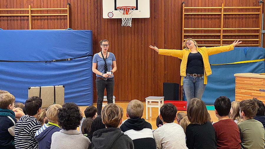 Zwei Lehrerinnen stehen vor einer Gruppe Schülerinnen und Schülern in der Turnhalle.