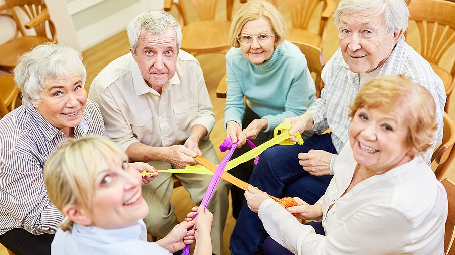 5 Seniorinnen und Senioren und eine Betreuerin sitzen im Kresi und machen eine Kräftigungsübung mit Gummibändern