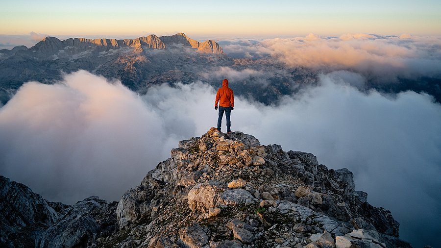 Mann steht mit dem Rücken zur Kamera auf einem Berggipfel und schau ins Tal